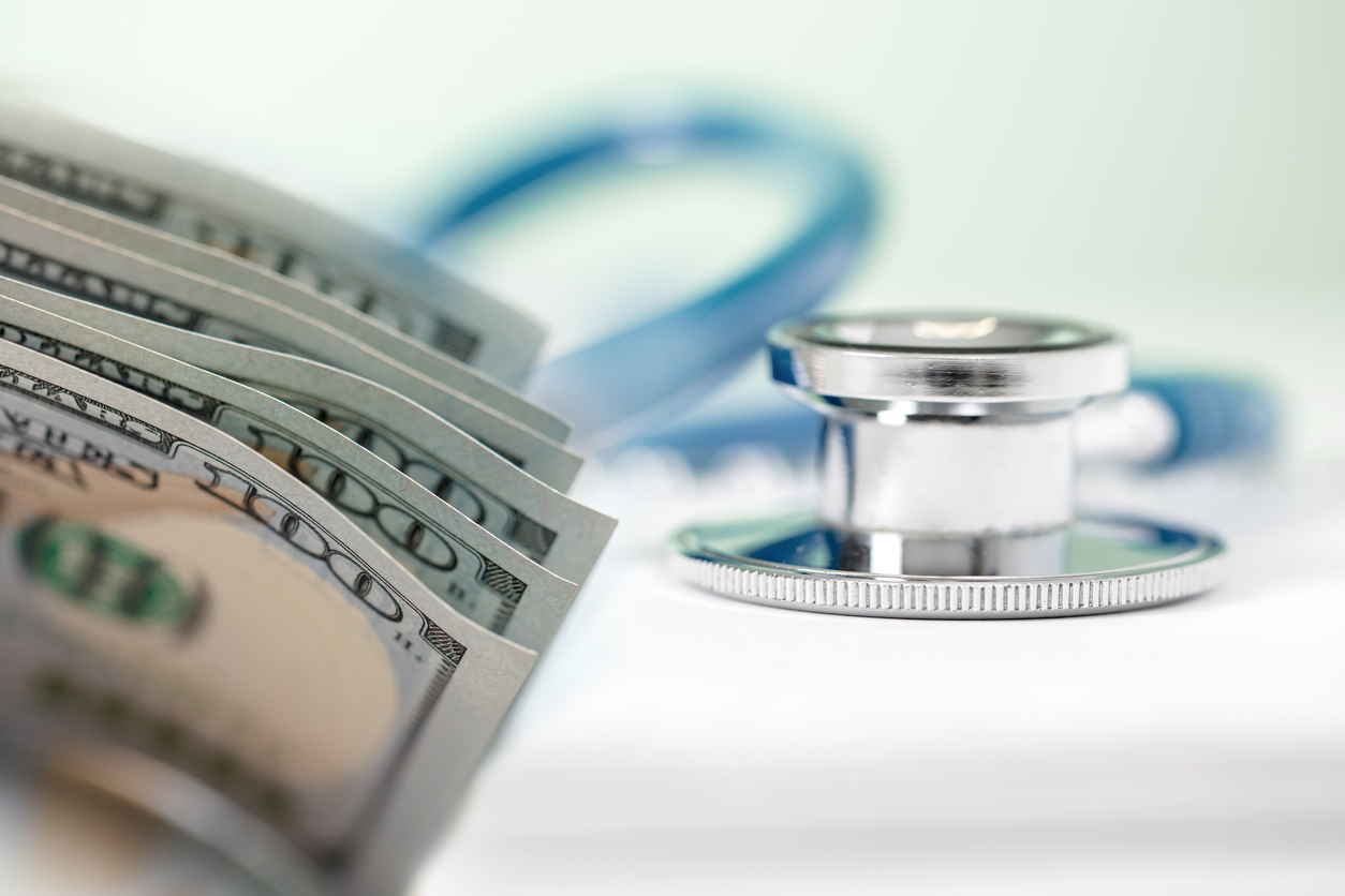 A roll of cash is the focus of the photo, with a stethoscope in the background.